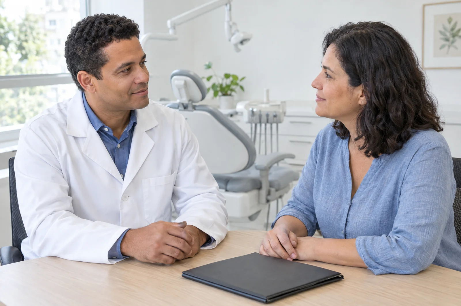 Cirurgião-dentista de jaleco branco conversa com uma paciente no consultório odontológico durante a avaliação.